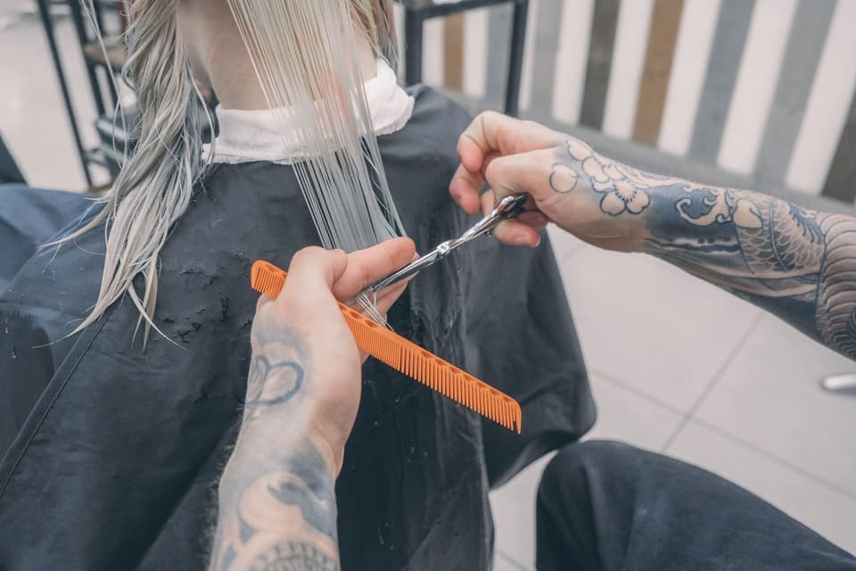 Stylist with tattooed hands cutting a client's blonde hair